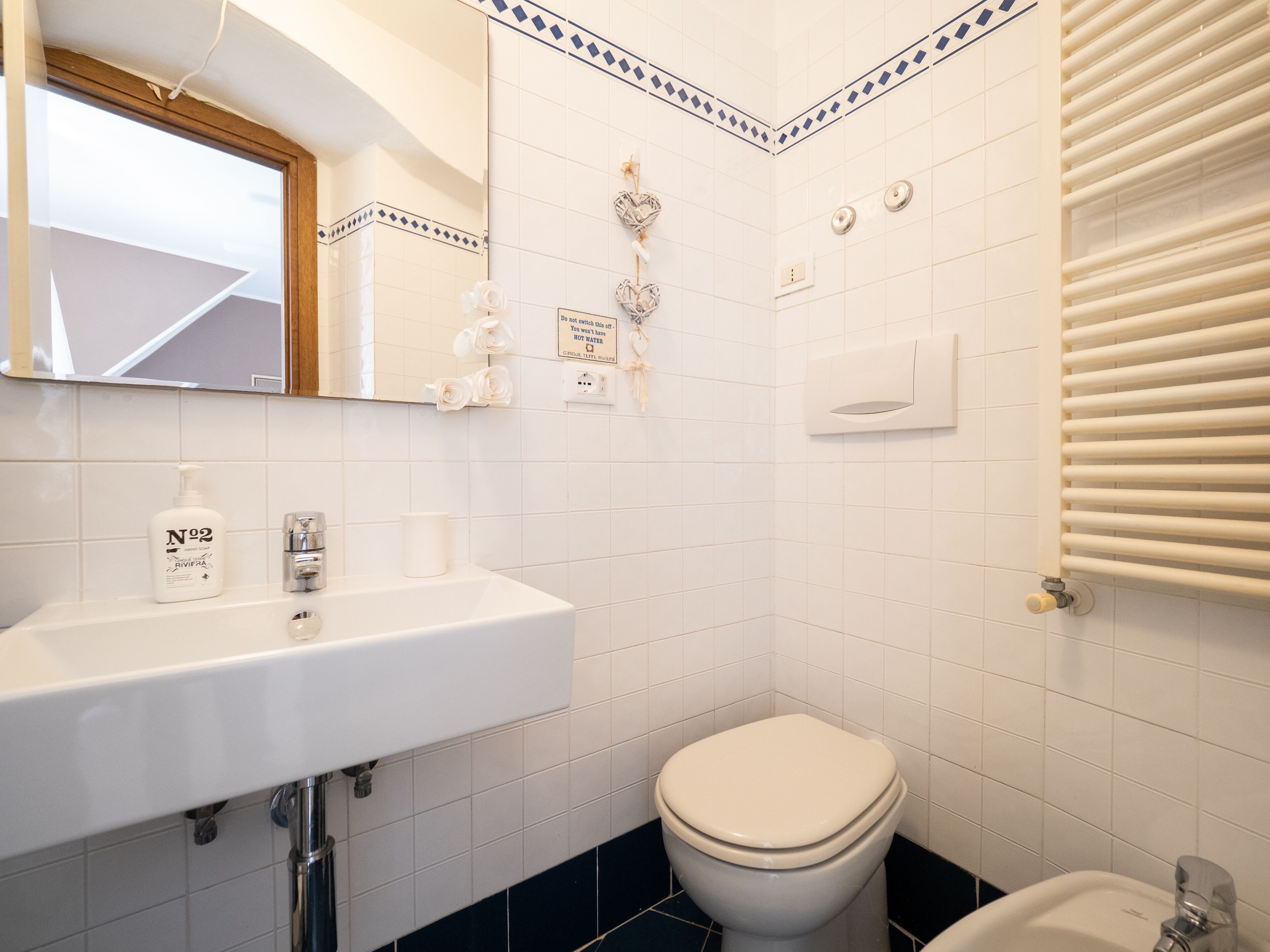 Une salle de bain avec un lavabo blanc, des toilettes et un miroir. On peut y voir des objets décoratifs accrochés au mur, notamment une décoration en forme de cœur et une pancarte indiquant : « Ne coupez pas l’eau. »
