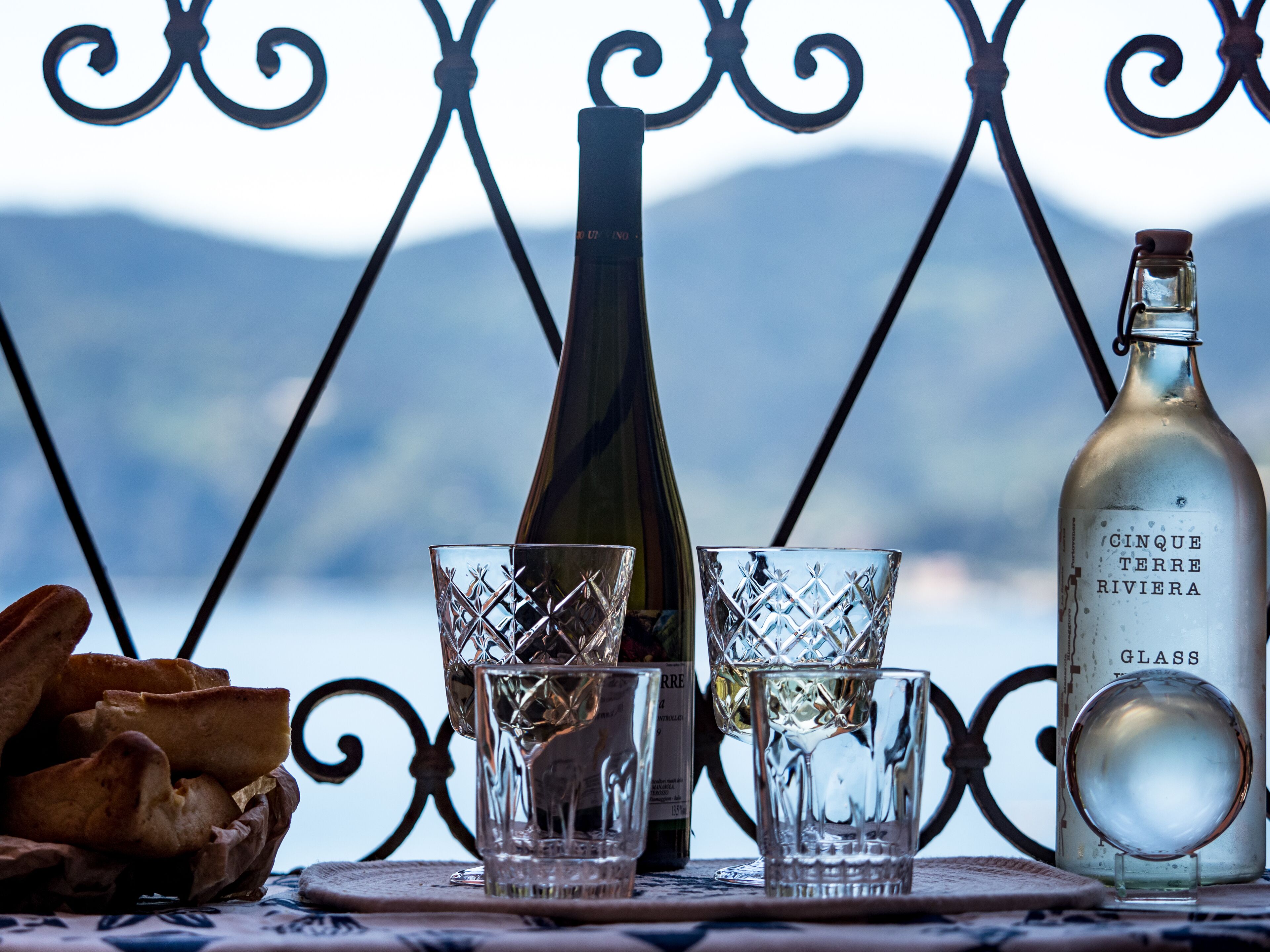 A table with a bottle of Cinque Terre Riviera wine, two glasses, and bread on a table with a metal railing in the background.