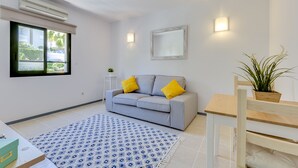 A modern living room with a grey sofa, yellow cushions, a blue patterned rug, a wooden dining table, and a potted plant.