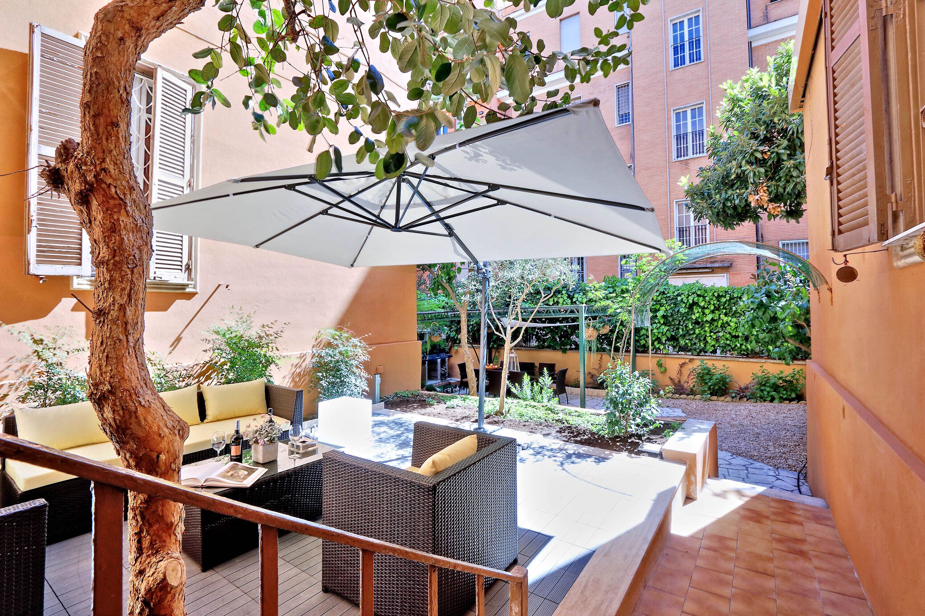Un patio al aire libre con mobiliario de mimbre, un paraguas blanco y el tronco de un árbol.