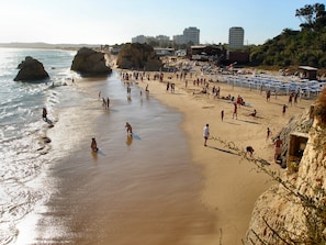 A crowded beach with people enjoying the sun and sea, rocky formations, and buildings in the background.