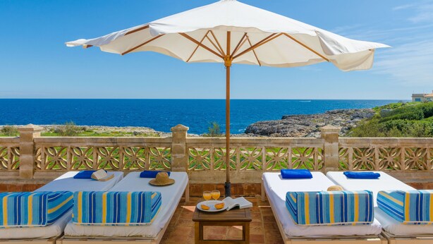 Eine Terrasse am Meer mit zwei Liegen, einem Tisch und einer Fruchtschale sowie einem großen Sonnenschirm.