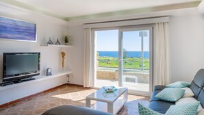 Ein Wohnzimmer mit Fernseher, Couchtisch und einer Couch mit gemusterten Kissen. Ein großes Fenster bietet Blick auf das Meer.