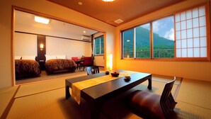 A traditional Japanese-style room with tatami flooring, a low table with cushions, and a view of a mountain through large windows. - Kuju Kanko Hotel (Kokonoe)