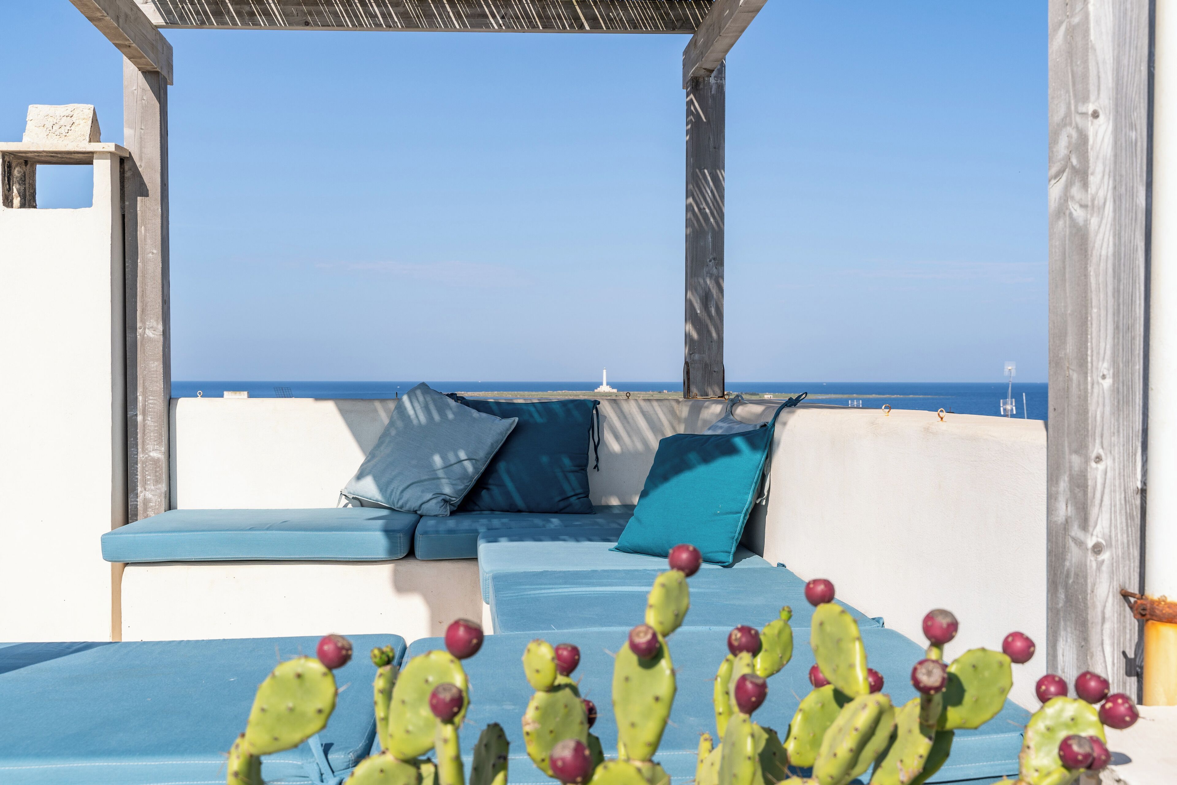 Um lounge no terraço com vista para o mar, com um sofá azul com almofadas e uma planta de cacto.