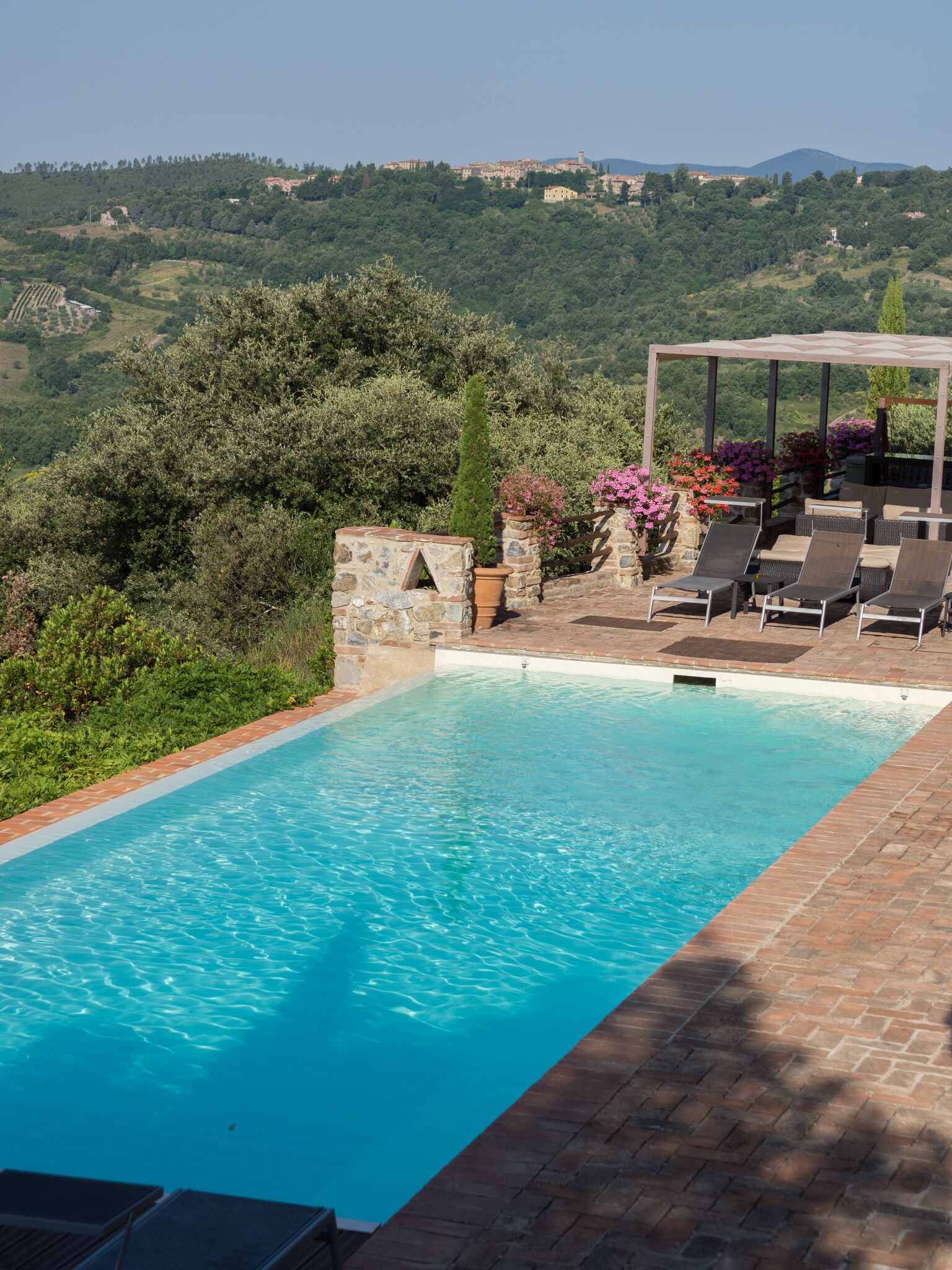 A swimming pool with a pergola and lounge chairs, surrounded by greenery and a view of a hilly landscape.