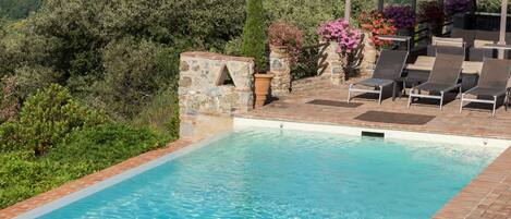 A swimming pool with a pergola and lounge chairs, surrounded by greenery and a view of a hilly landscape.