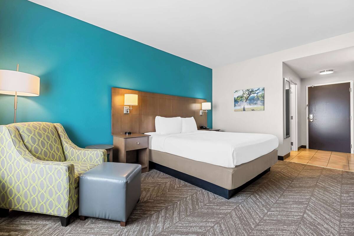 A hotel room with a bed, a nightstand, a lamp, a chair, and a small ottoman.