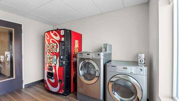 Une buanderie équipée d’un distributeur automatique rouge et noir, d’un lave-linge argenté et d’un sèche-linge.