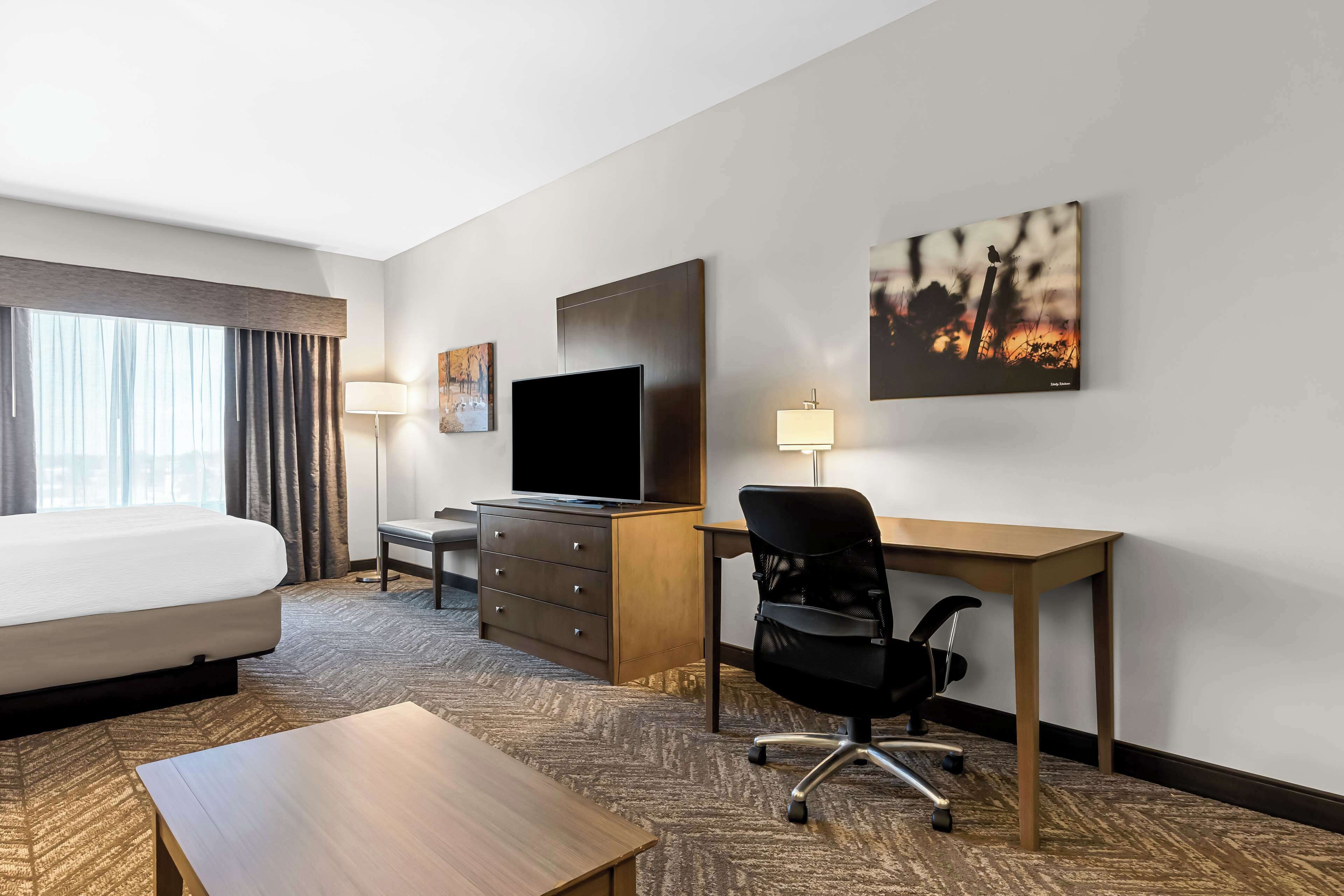 A hotel room with a bed, desk, chair, TV, and artwork on the wall.
