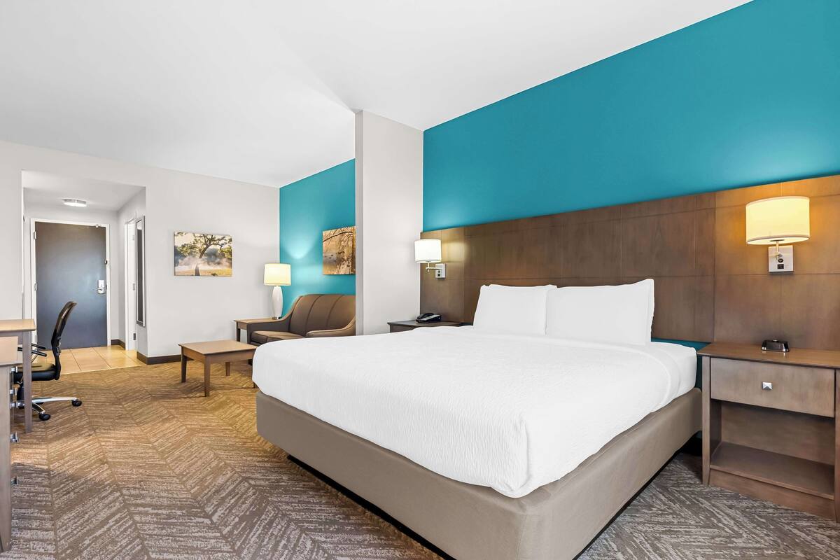 A hotel room with a large bed, two bedside tables, a desk, a chair, and a door leading to another room.