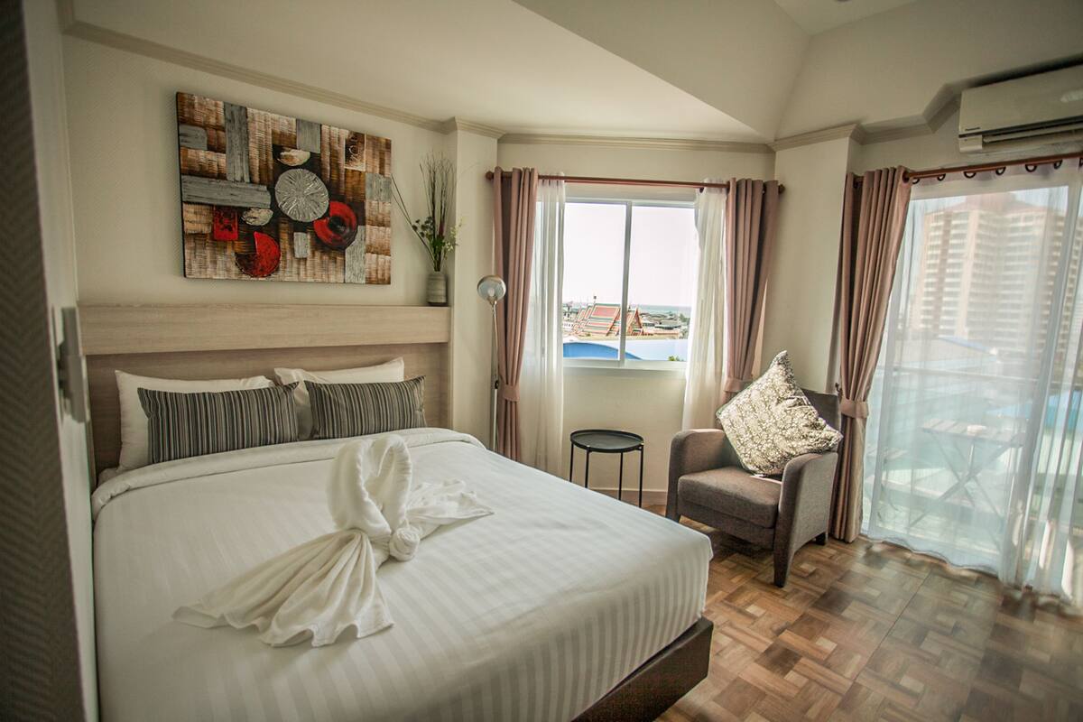 A hotel room with a large bed, a chair, a small table, and a view of the city through the window.
