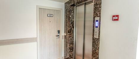 Un ascensor moderno con pantalla digital, una puerta marcada con el número 507 y un enchufe de emergencia rojo en la pared.
