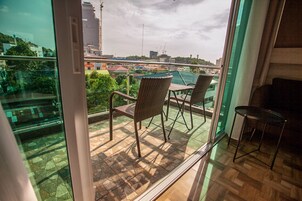 Un balcón con sillas de mimbre y una mesita, con vistas a un paisaje urbano con edificios y zonas verdes.