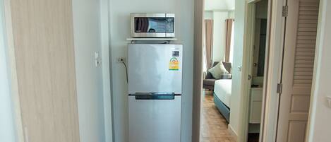Un pasillo estrecho con un refrigerador, suelo de madera y una puerta que da a un dormitorio con cama y cortinas.