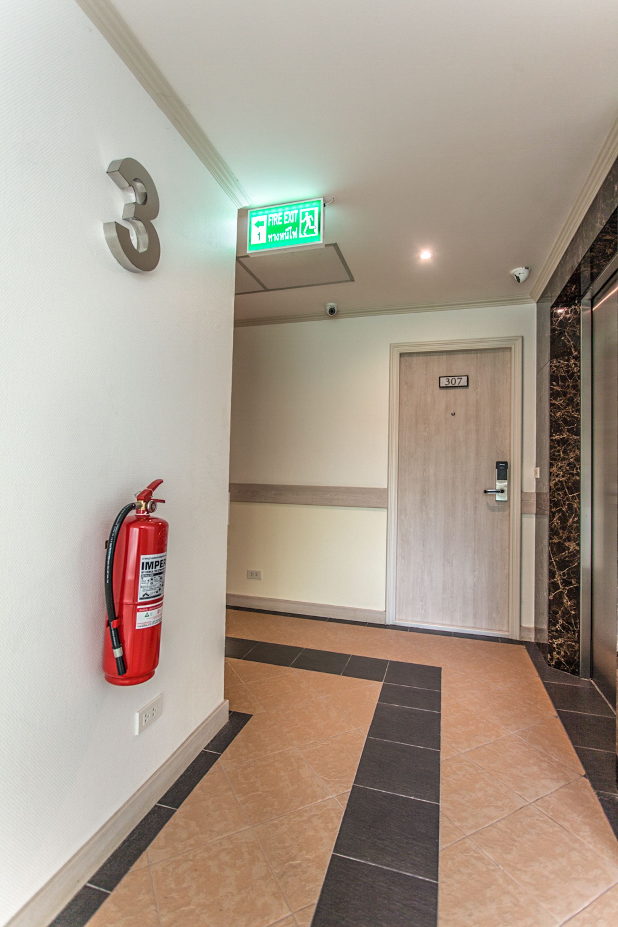 A hallway with a fire extinguisher mounted on the wall, a door labeled "307", and a green exit sign above the door.
