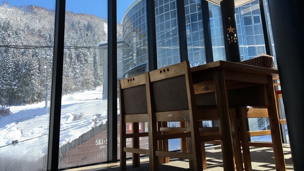 A room with large windows offering a view of a snowy landscape and a building with a glass facade. - Lodge Nakajima (Nozawaonsen)