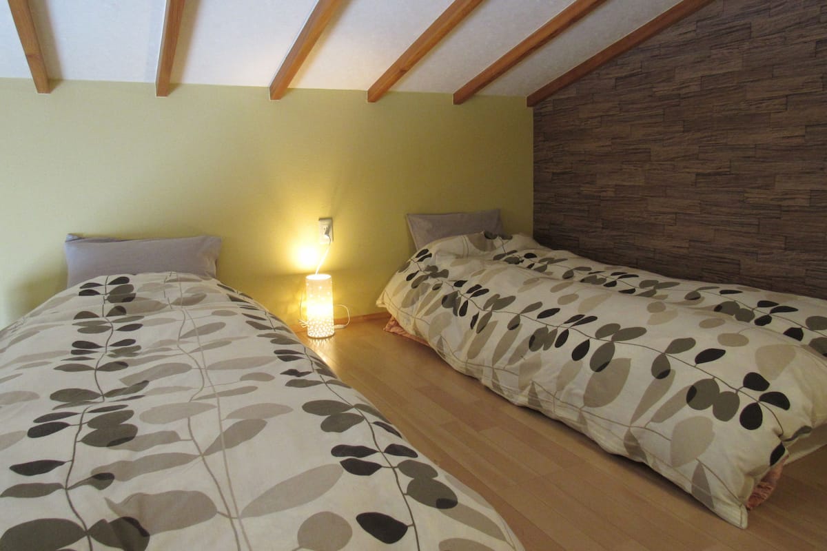 A bedroom with two single beds, wooden beams on the ceiling, a wall-mounted lamp, and a wooden textured wall.