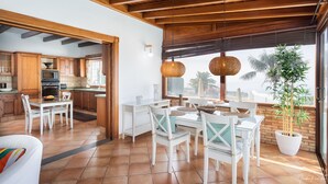 Une cuisine spacieuse avec des meubles en bois, un coin repas avec des chaises blanches et une grande fenêtre donnant sur des palmiers.