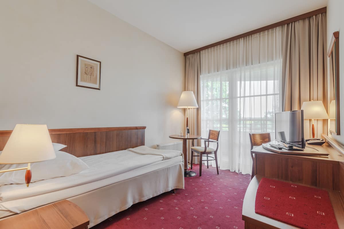 Ein Hotelzimmer mit Bett, Nachttisch, Lampe, Stuhl, Schreibtisch mit Computer und einem Fenster mit Vorhängen.