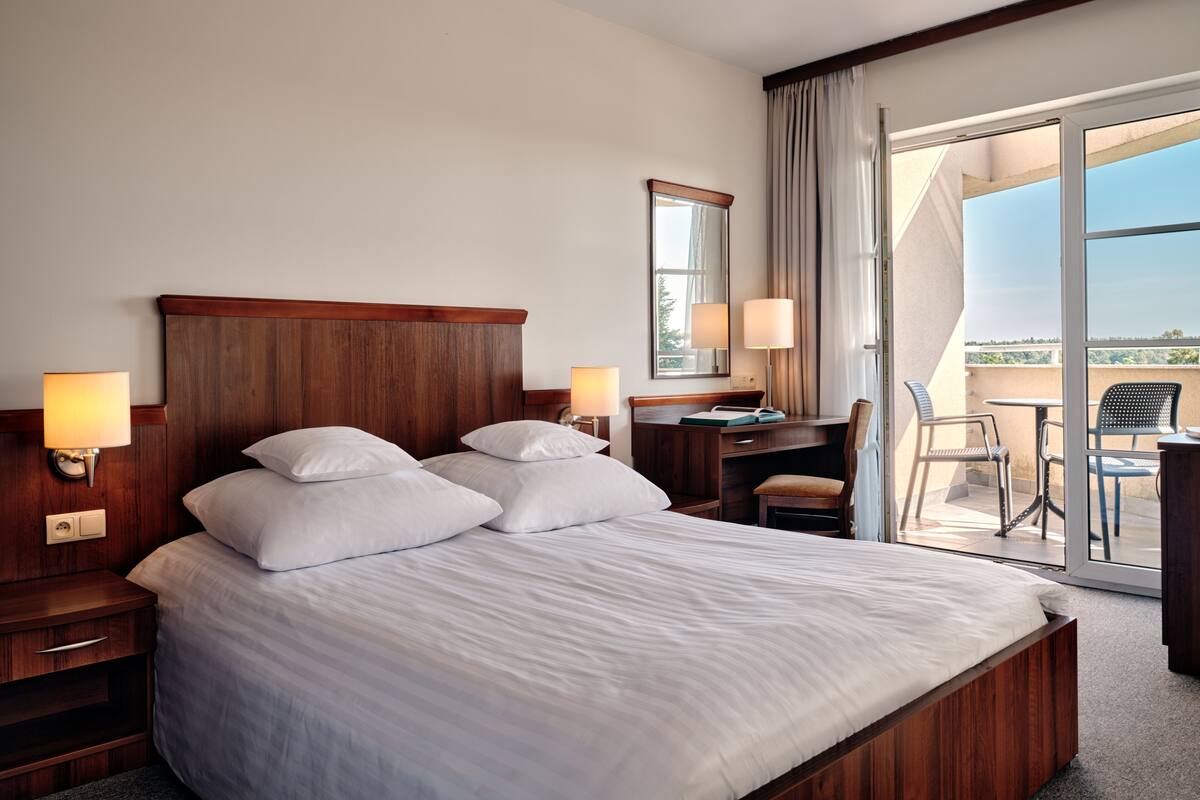 Ein Hotelzimmer mit einem großen Bett, Nachttischlampen, einem Schreibtisch mit Stuhl und Blick ins Freie durch eine Schiebetür.