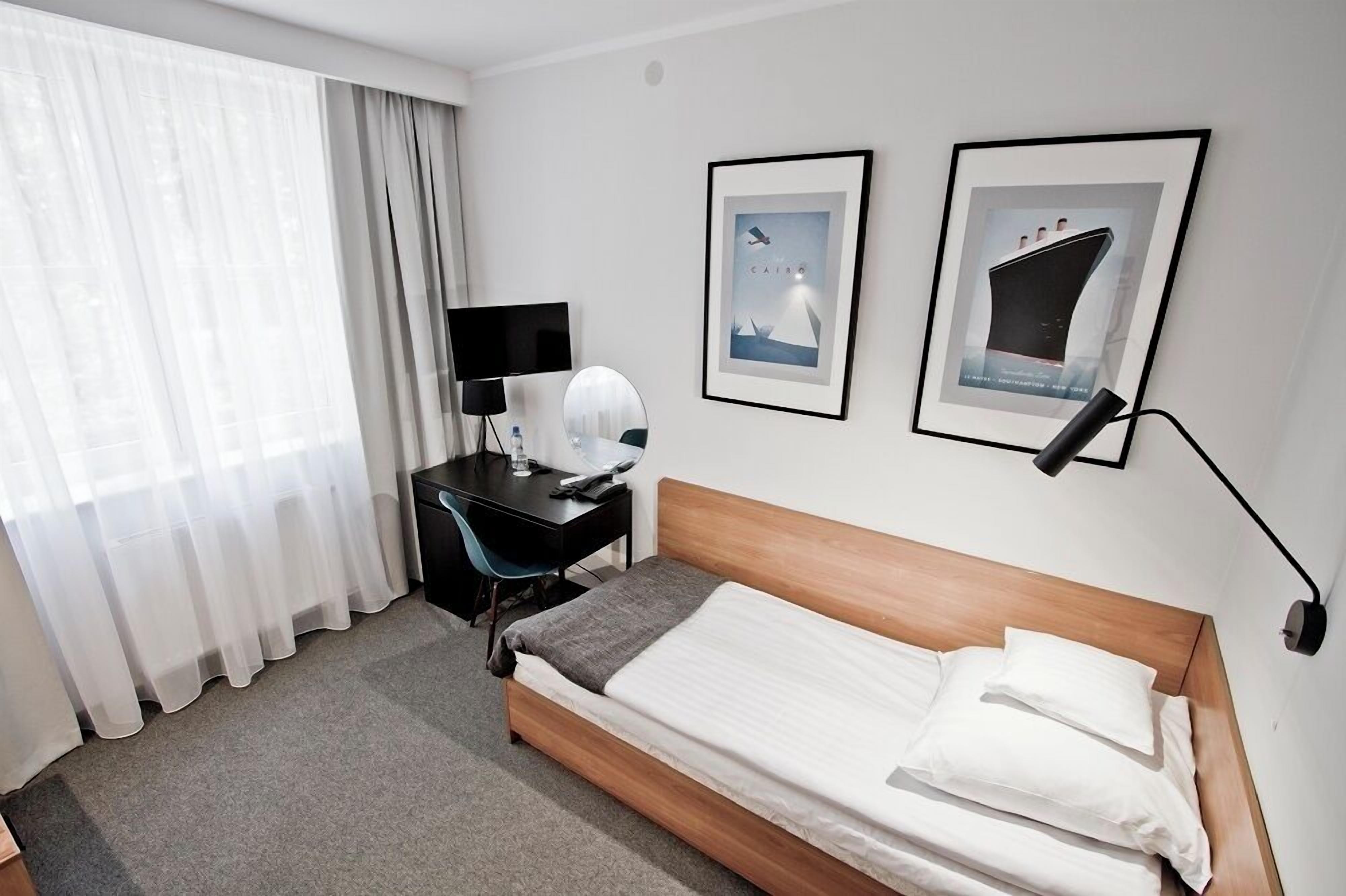 A hotel room with a bed, a desk, a chair, a TV, and two framed pictures on the wall.