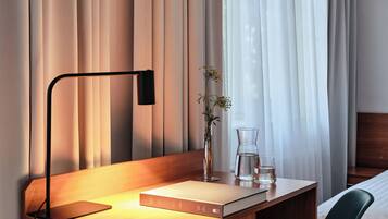A hotel room with a wooden desk, a modern chair, a bedside table with a glass of water, a vase with flowers, and a bed with a wooden headboard.
