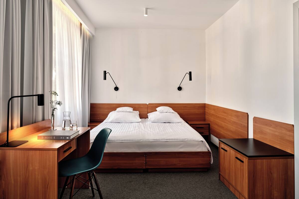 Ein Hotelzimmer mit einem hölzernen Bett, einem Schreibtisch mit Stuhl, einem Nachttisch und zwei Wandleuchten.