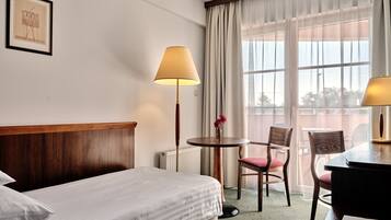 Ein Hotelzimmer mit Bett, Schreibtisch, Stuhl, Lampe, einem Fenster mit Vorhängen und einem gerahmten Bild an der Wand.