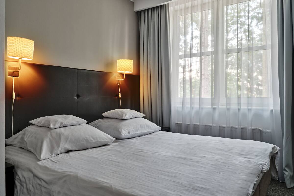 Ein Hotelzimmer mit einem großen Bett, zwei Nachttischlampen, einem Fenster mit durchsichtigen Vorhängen und einer Heizung.