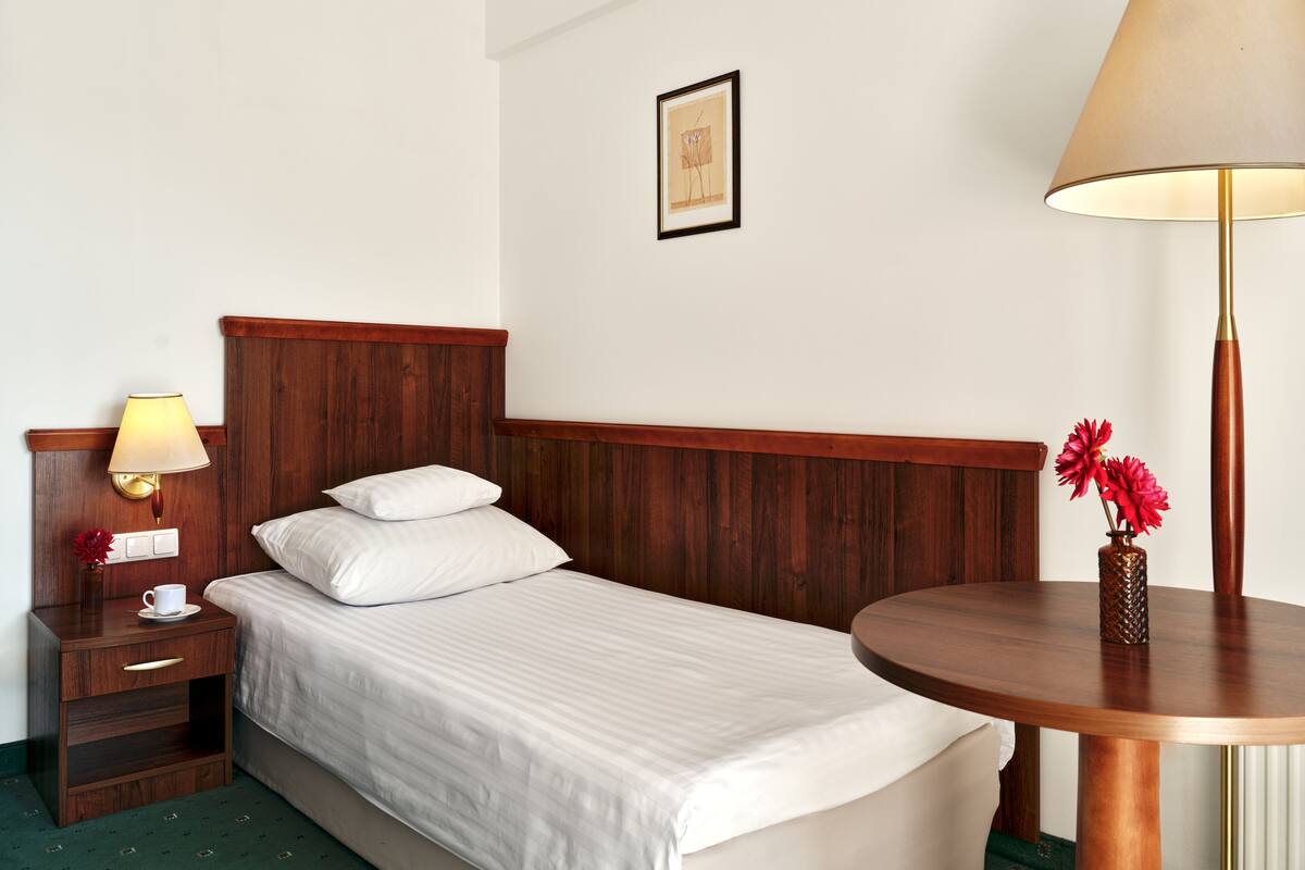 Ein Hotelzimmer mit einem Bett mit Holz-Kopfteil, einem Nachttisch mit Lampe, einem kleinen Tisch mit Blumenvase und einem gerahmten Bild an der Wand.