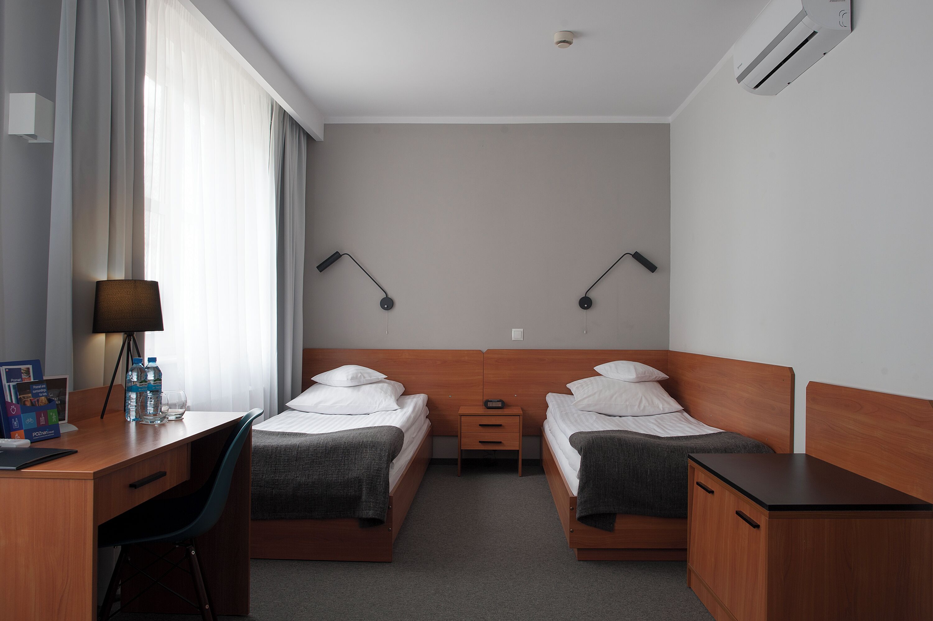 Camera d'albergo con due letti singoli, una scrivania con sedia, un piccolo comodino e una lampada.