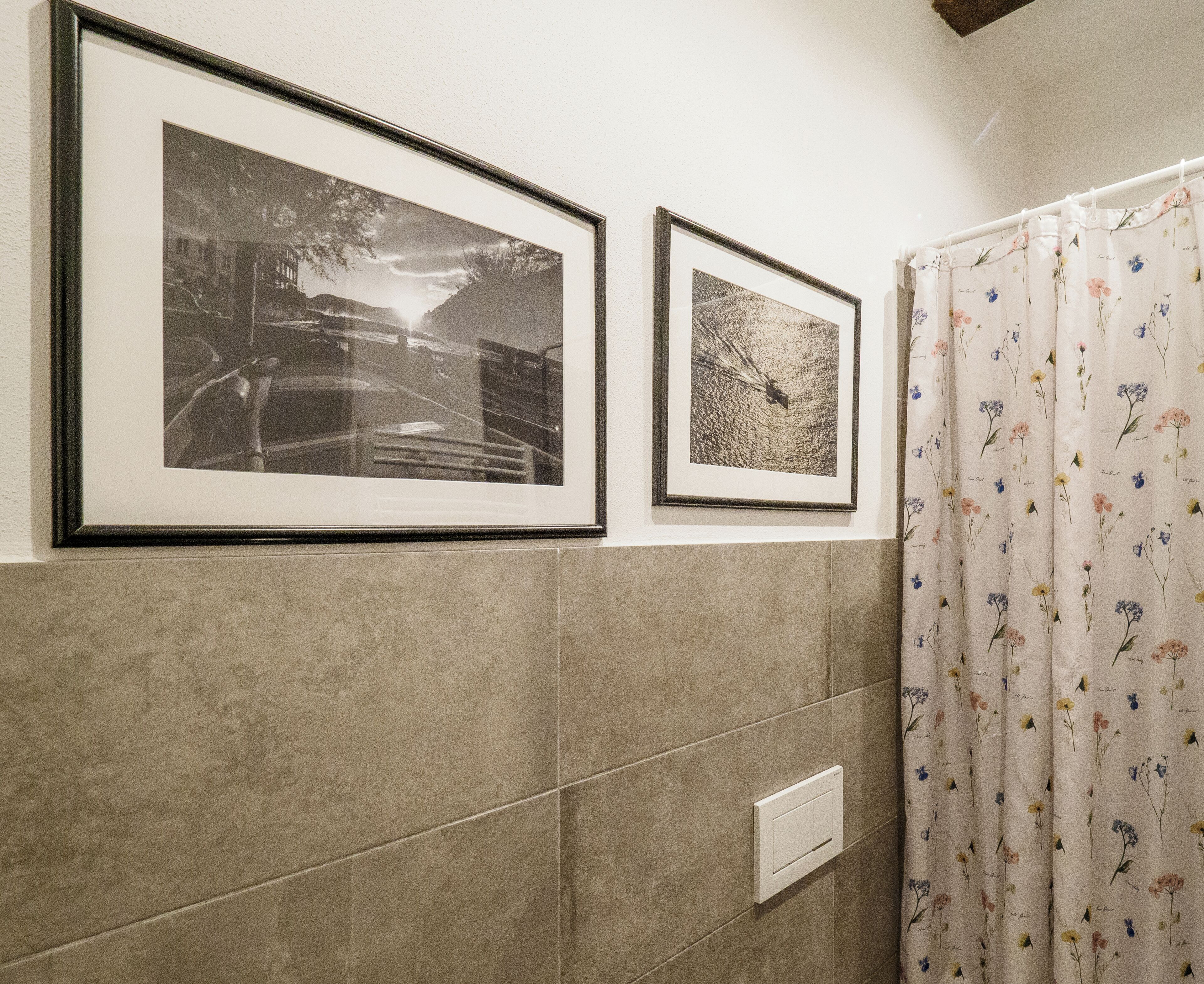 Un baño con dos fotografías enmarcadas en la pared, una cortina de ducha con motivos florales y un interruptor de luz.