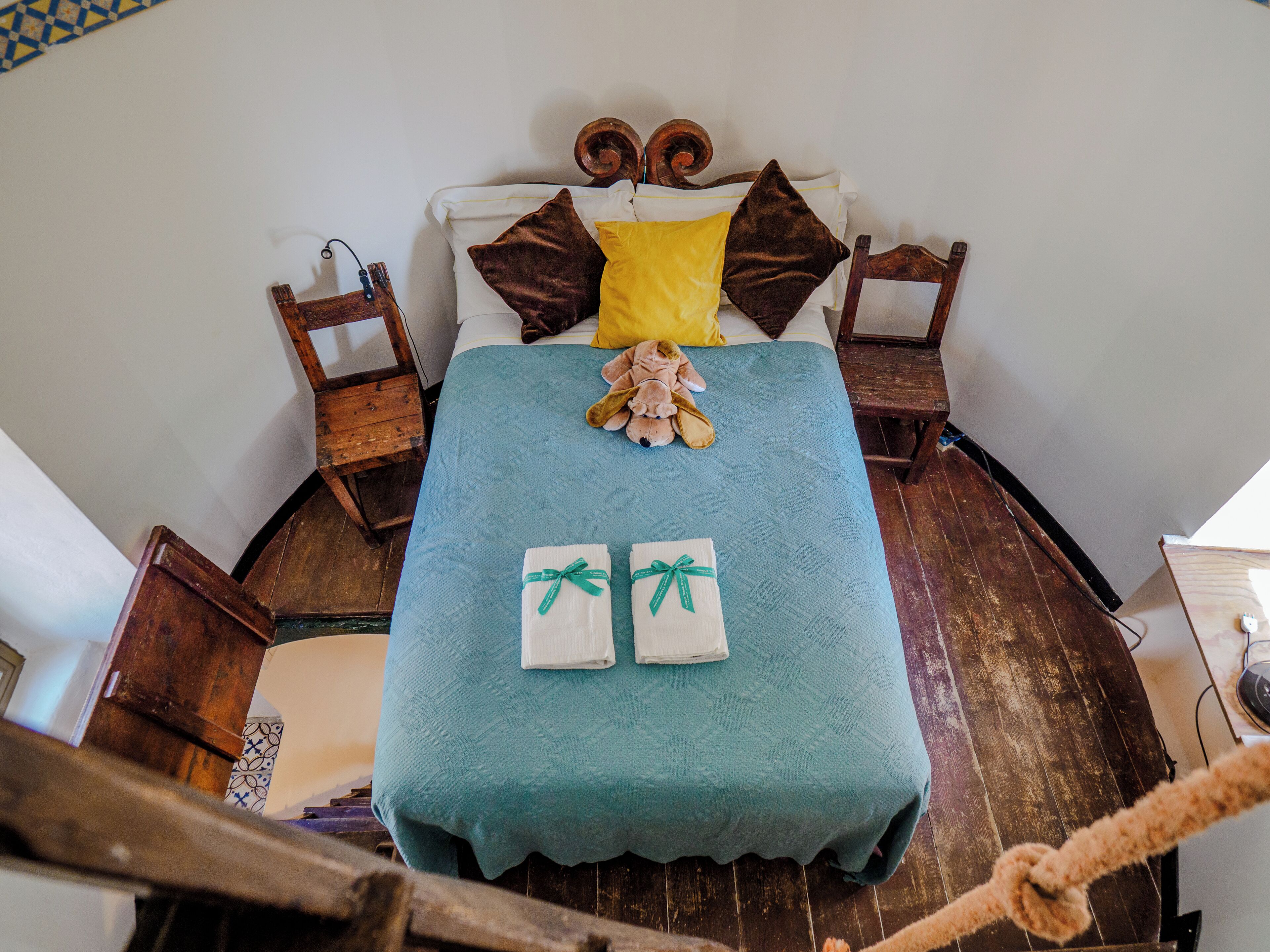 A bed with a teal bedspread, two pillows, a teddy bear, and two towels.