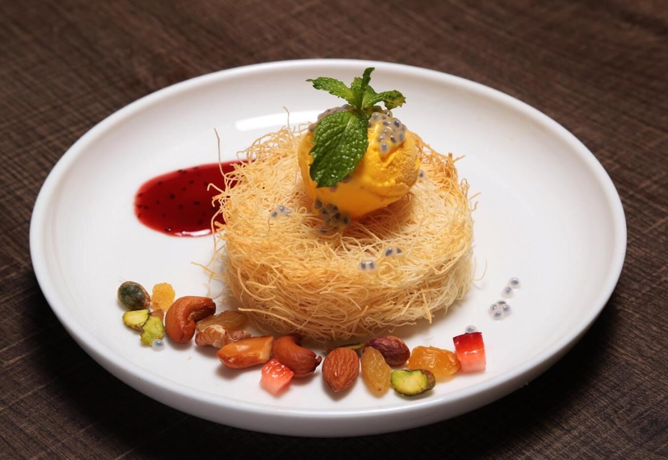 A dessert on a white plate with a red sauce, a mint leaf, and various nuts.