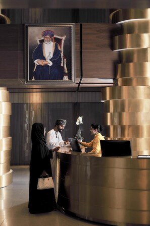 A reception area with a reception desk, a framed portrait above the desk, and a modern, curved metallic column structure. - Aerotel Muscat - Airport Transit Hotel (Muscat)