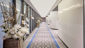 Un pasillo moderno de hotel con alfombra estampada, ventanas amplias y arreglos florales decorativos.