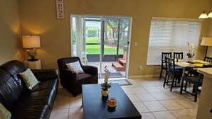 A living room with a black leather sofa, a dark armchair, a coffee table with a plant, a dining area with a table and chairs, and a sliding glass door leading to a patio with a lawn and a fence.