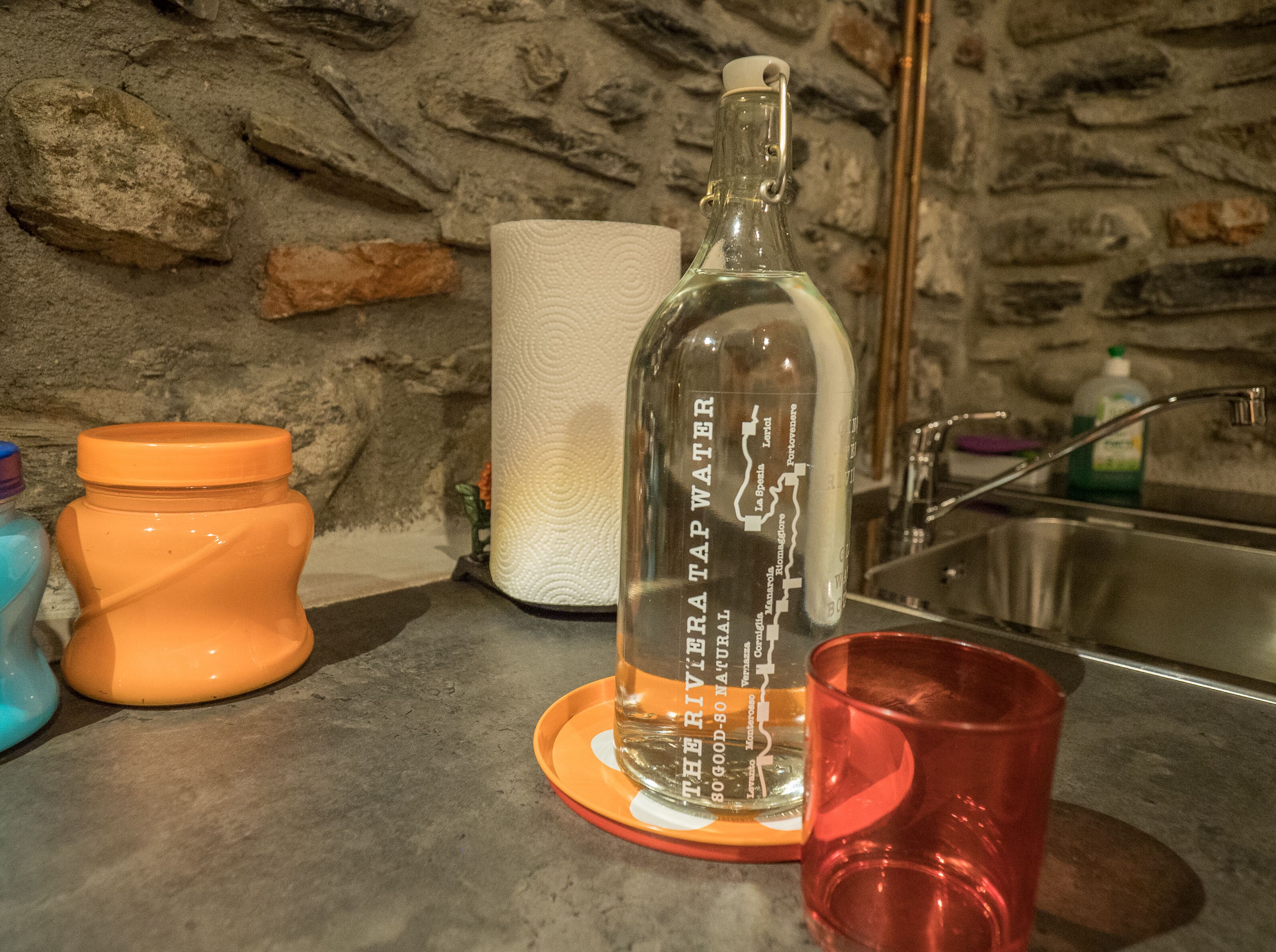 En köksbänk med en flaska kranvatten, en rulle hushållspapper, en glasburk och ett rött glas.