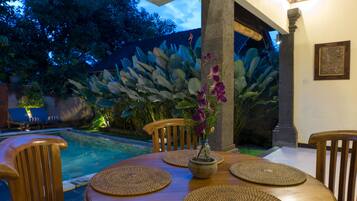 A wooden dining table with woven placemats, a vase with flowers, and a view of a swimming pool and tropical plants.