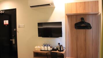 A hotel room with a flat-screen TV mounted on the wall, a wooden desk with a chair, a hanging coat rack, and a small shelf with toiletries.
