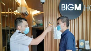 A man in a white shirt conducting a temperature check on another man in a blue shirt at a hotel reception area with a sign reading "BIGM HOTEL".