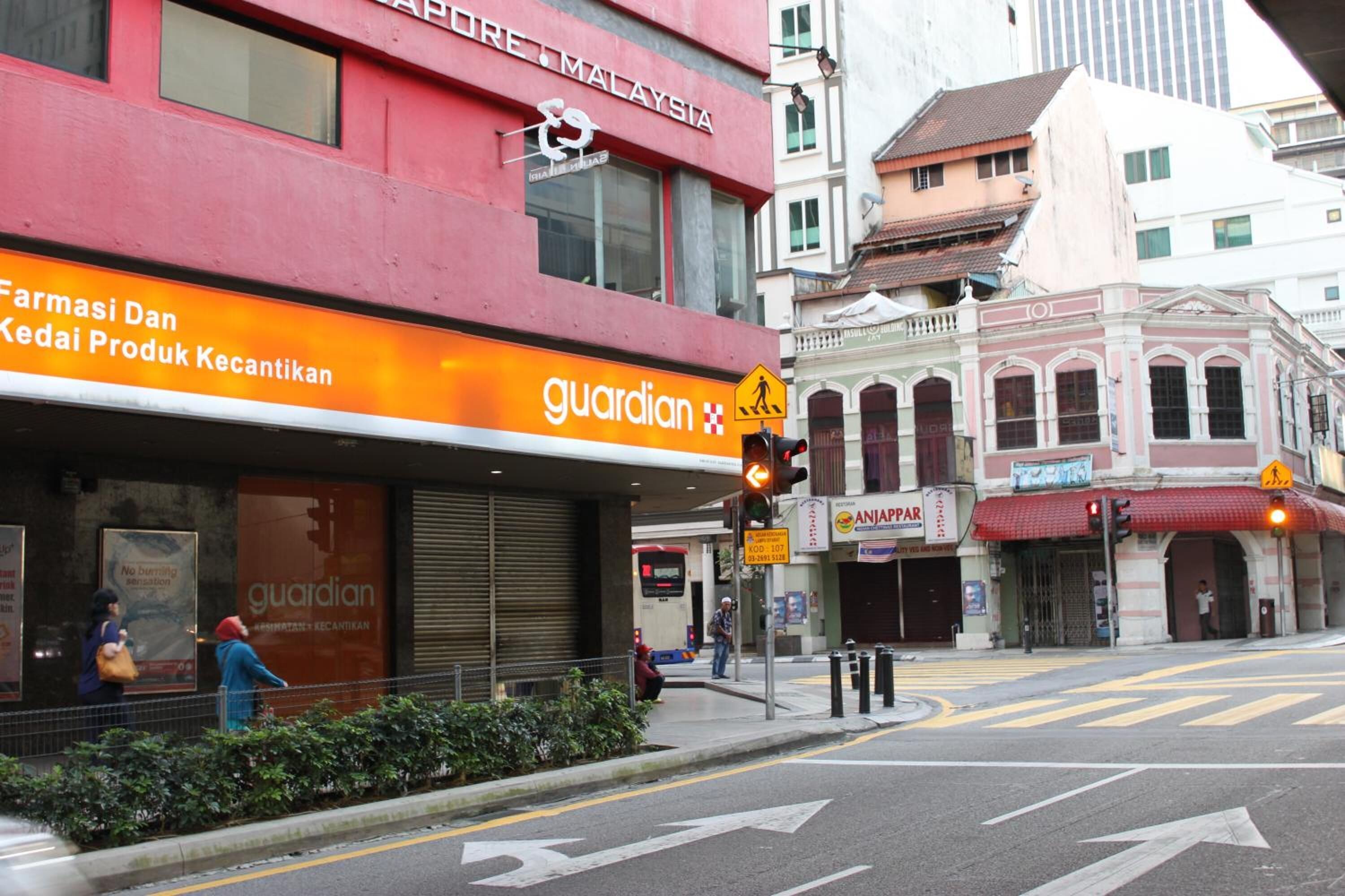 Vista de la calle con una tienda Guardian, un edificio rojo con el rótulo 'APURE MALAYSIA', y un edificio tradicional con toldo rojo.