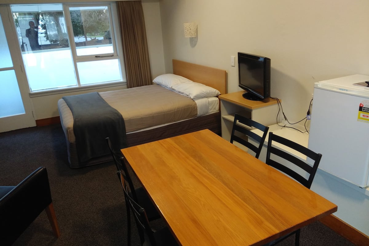 Een kleine hotelkamer met een enkel bed, een bureau met een televisie, een koelkast en een raam met gordijnen.