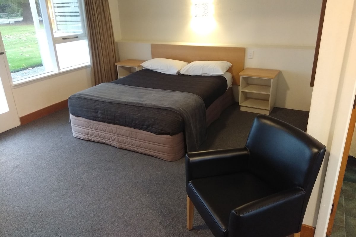 Een hotelkamer met een bed, twee nachtkastjes, een stoel en een raam met gordijnen.