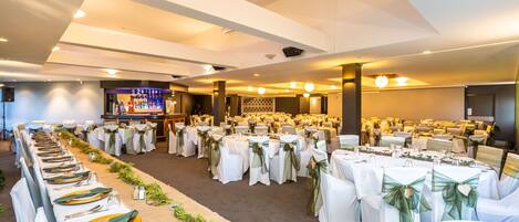 Een feestzaal met lange tafels gedekt voor een formeel evenement, met witte tafelkleden en groene linten.