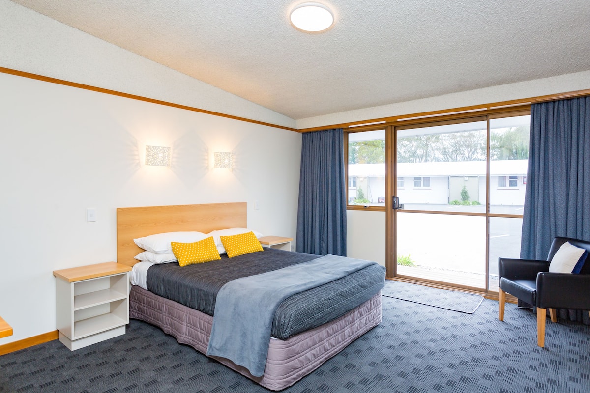 Een hotelkamer met een groot bed, een nachtkastje, een stoel en een raam met gordijnen.