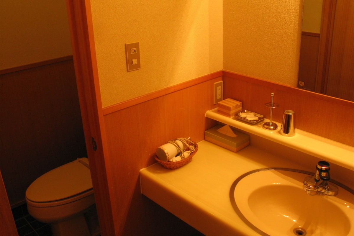 A bathroom with a toilet, a sink, and a mirror. There are toiletries on the sink and a basket on the floor.