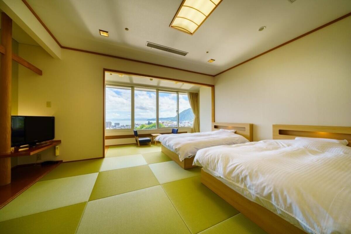 Une chambre d’hôtel avec deux lits, une télévision, une grande fenêtre donnant sur un beau paysage et un sol recouvert de tatamis.