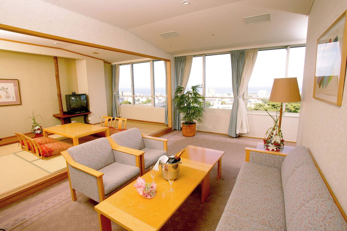 Habitación de hotel con sofá, sillas, una mesita, televisor, planta en maceta y vista a la ciudad.